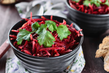 Thinly sliced ​​fresh beets in a black salad bowl with sprigs of cilantro ready for eating are on a wooden table. Shredded beet salad, closeup.