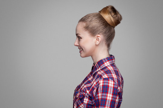 profile side view of beautiful blonde girl in red, pink checkered shirt, collected bun hairstyle standing and looking aside with toothy smile. indoor studio shot. isolated on gray background