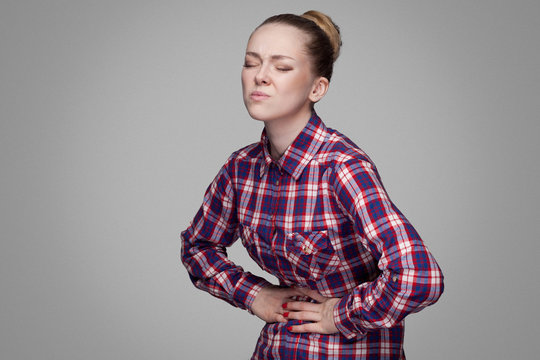 stomack sick unhappy girl in red, pink checkered shirt, collected bun hairstyle, makeup standing and touching her painful belly. indoor studio shot. isolated on gray background