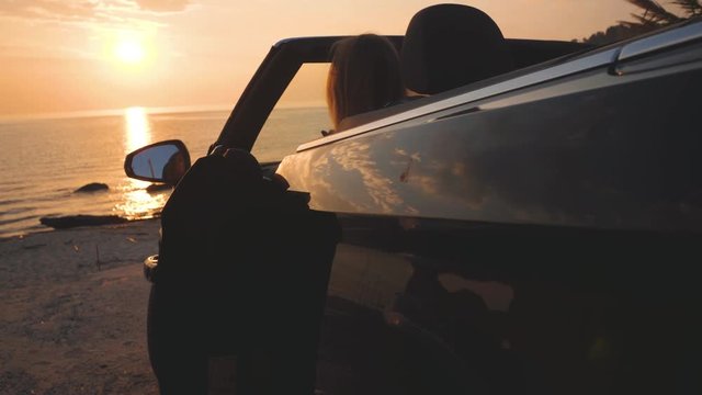 Pretty girl in dress goes out of luxury car and runs towards sea and sunset sky