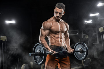 Young handsome sexy man, athlete, bodybuilder, weightlifter, in a modern gym is covered with a dark background, doing exercises for the biceps using sporting goods - weights.  - 242043563