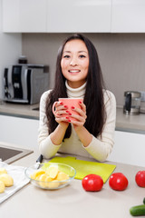 Image of young brunette with mug in hands standing at table with vegetables and fruits