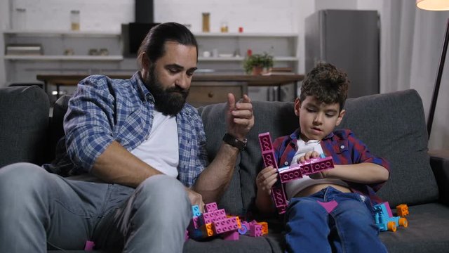 Cheerful single father and joyful boy creating toys from colored plastic blocks while relaxing on sofa in domestic room. Caring dad with son busy playing with toy construction bricks at home.