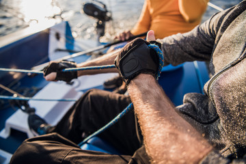 Close-up of male hands in gloves pulling rope of sailboat while mooring yacht and sitting with friends on deck.
