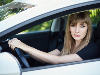 Happy young woman posing in her car holding steering wheel. Lady driver with long blond healthy hair at vehicle. Warm season lifestyle portrait.