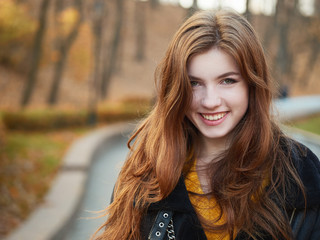 Fabulous redhead woman with long hair in yellow sweater black leather jacket on blurred autumn background.