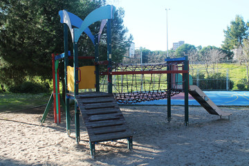 Playground for children, located in the park, consisting of a swing and suspension bridge. Colors red, green, blue and yellow. Located on sand. Image taken during day.