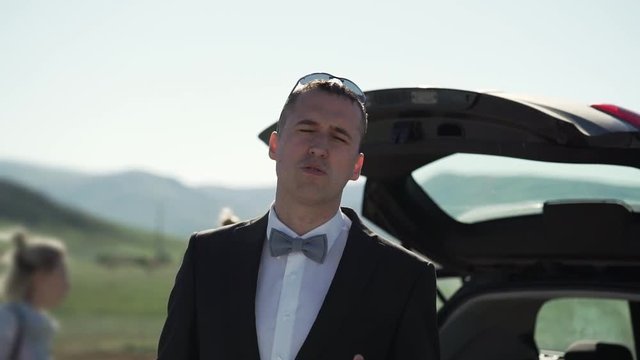A young man in a classic suit, bow tie smokes VAPE. Stands next to the car in nature.