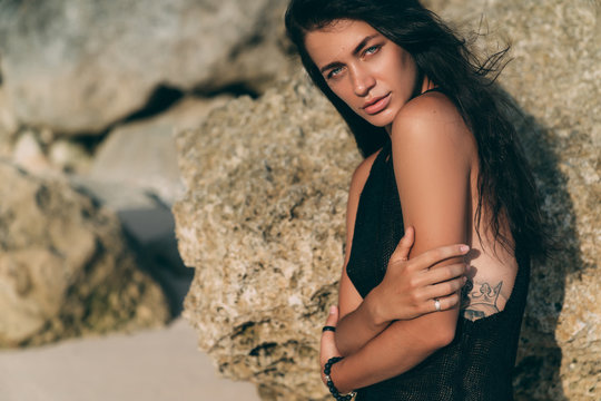 Portrait of sexy brunette model in knitted sweater posing on the sandy beach, big stones and rock on background.