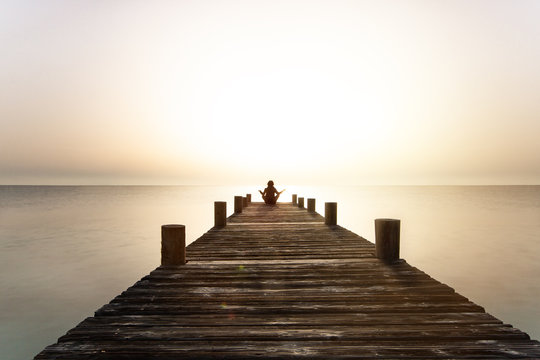 Person meditiert bei Sonnenuntergang auf einem Steg am Meer
