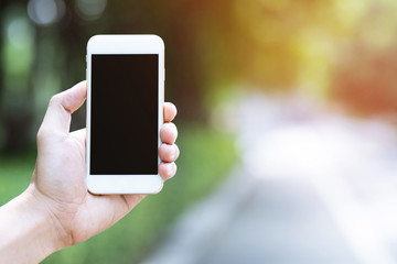 close up people hand holding display white mobile smart phone with blank or empty black screen on outdoor public parks green tree background. soft focus.
