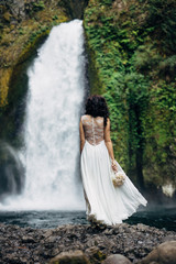 bride in wedding dress standing near large waterfall holding bouquet