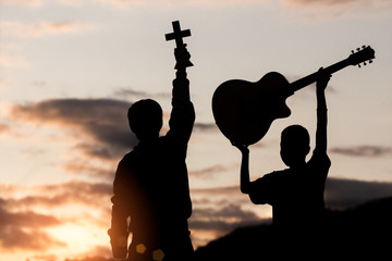 Two young christian worship God by holding cross and guitar on moutain with light sunset...