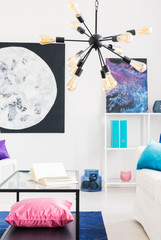 Vertical view of black modern chandelier with yellow bulbs in cosmos inspired living room with moon and galaxy graphic and metal coffee table with pink pillow on it, real photo