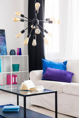 Black modern chandelier with yellow bulbs above metal and glass coffee table with book on it in bright living room with white sofa with pillows, real photo