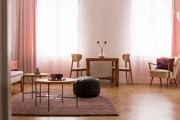 Wooden table and pouf on carpet in living room interior with chairs, armchair and windows. Real photo