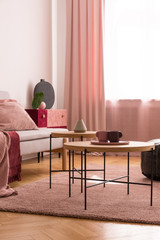 Wooden tables on pink carpet in living room interior with grey settee and drapes. Real photo
