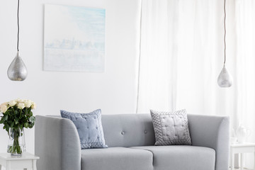 Pillows on grey sofa in white living room interior with lamps, flowers and poster. Real photo