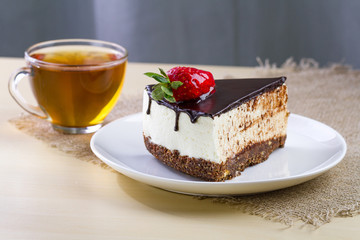 A cup of hot tea and a slice of sweet cake with fresh strawberries and dripping chocolate glaze in a white plate. Sweet dessert