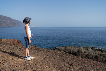 A boy in shorts, a T-shirt and a hat is standing on a rocky seashore and looking into the distance.