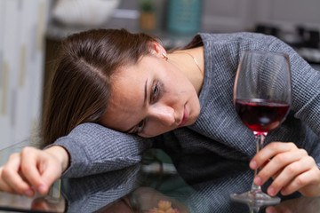 Depressed, divorced crying woman sitting alone in kitchen at home and drinking a glass of red wine because of problems at work and troubles in relationships. Social and life problems