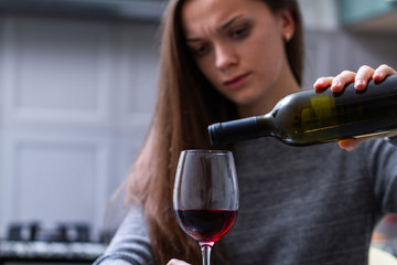 Depressed, divorced crying woman sitting alone in kitchen at home and drinking a glass of red wine because of problems at work and troubles in relationships. Social and life problems