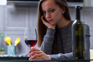 Depressed, divorced crying woman sitting alone in kitchen at home and drinking a glass of red wine because of problems at work and troubles in relationships. Social and life problems