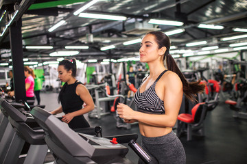 Nice young woman runs on running machine. She looks concentrated. Young asian woman runs as well further. They in gym room alone.