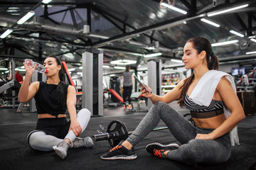 Fototapeta na wymiar Two young sportwomen sit on floor on gym. They have rest. Asian model drnk water. European hold phone and look at it. She smiles.