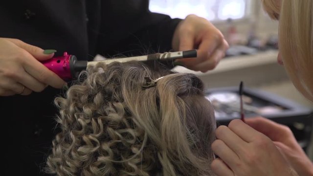 Professional hairdresser stylist makes curling light brown hair by tongs. Makeup artist makes makeup.