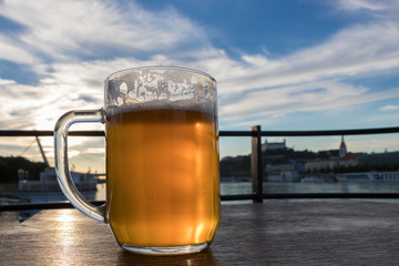 Close-up of one cold beer mug with foam and water drops on the background blue sky and white clouds and the sun