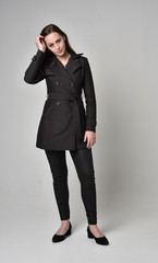 full length portrait of a brunette girl wearing long black coat, standing pose on grey studio background.
