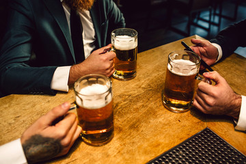 Picture of mn in suits sit at table. They hold mugs of beer and one phone.