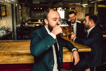 Young man in suit stand in bar and drink beer. Two other men stand behind him. They talk.
