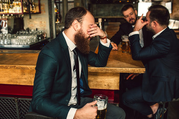 Tired young man sit and cover face with hand. Two other men sit behind and talk. They are in bar.