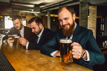 Cheerful and positive bearded man in suit look on camera. He smile and hold beer mug. Other two young men sit behind.