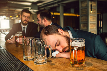 Tired young man in suit sleep on bar counter. He is drunk. there are two empty mugs and one full...