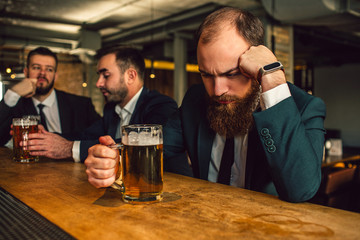 Young man in suit sit and sleep. He prop up head. Guy hold beer mug. Other two office workers sit behind and talk.