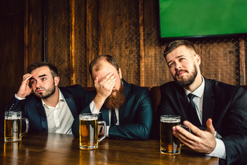 Three young office workers in suits sit at table in bar. They watch football game. Guy on middle cover face with hand. They all are emotional.