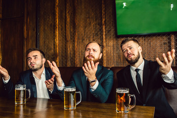 Emotional and upset young men in suits sit at table in bar. They wave with hands and look forward. Guys watch football game.