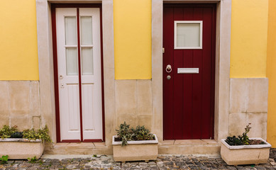 Fototapeta na wymiar Two ancient wooden doors in Lisbon, Portugal. White and red doors with big flower pots. Traditional portuguese exterior. Entrance concept. Old european architecture. Yellow wall and colorful doors.