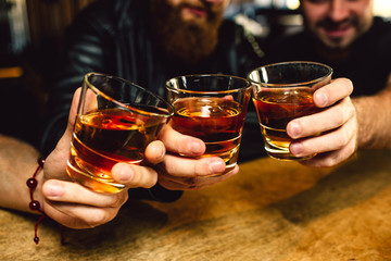 Cut view of three bearded young men holding glasses with rum together. They smile. People sit in...