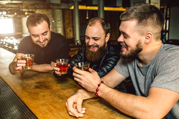 Three friends sit together at bar counter. They hold glasses of alcohol in hands. Men smile.
