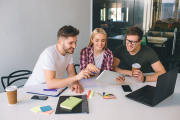 Fototapeta premium Young woman and two men sit at table in bright room. They look at tablet and smile. Cup of coffee, notebook and stickers are on table