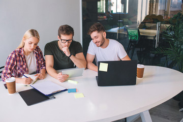 Three coworkers sit at table. Young woman look serious. She write in notebook. Guy in black shirt...