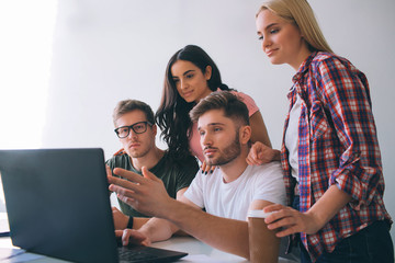 Serious young men sit at table and look at laptop screen. Brunette and blone young women stand...