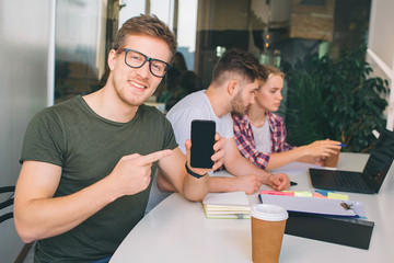 Nice young man in glasses point on hpone in hands. He look on camera. Other two young people work...