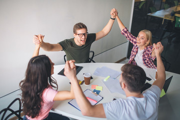 Happy young man and woman hold each other hands up and joying. They sit around white table in room. Team rejoicing.