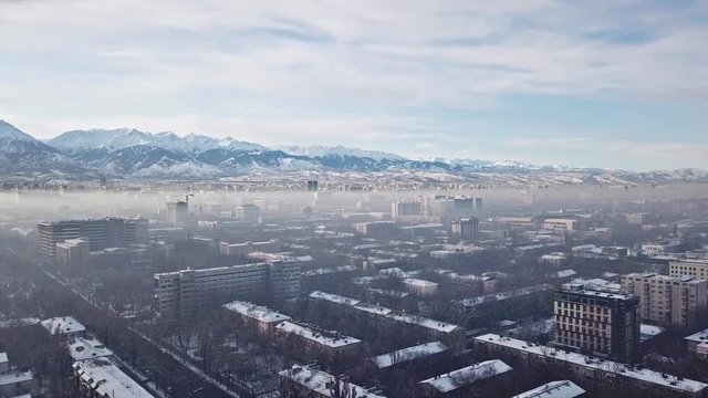 Shooting from a drone over the big city of Almaty. View of many roads, cars, people and large mountains. Places smog and fog. People walk the streets. Beautiful buildings with glass Windows. Trees.