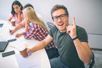 Cheerful nice young man in glasses looks on camera. He rocks. Other three young people sit behind hm and work in team.